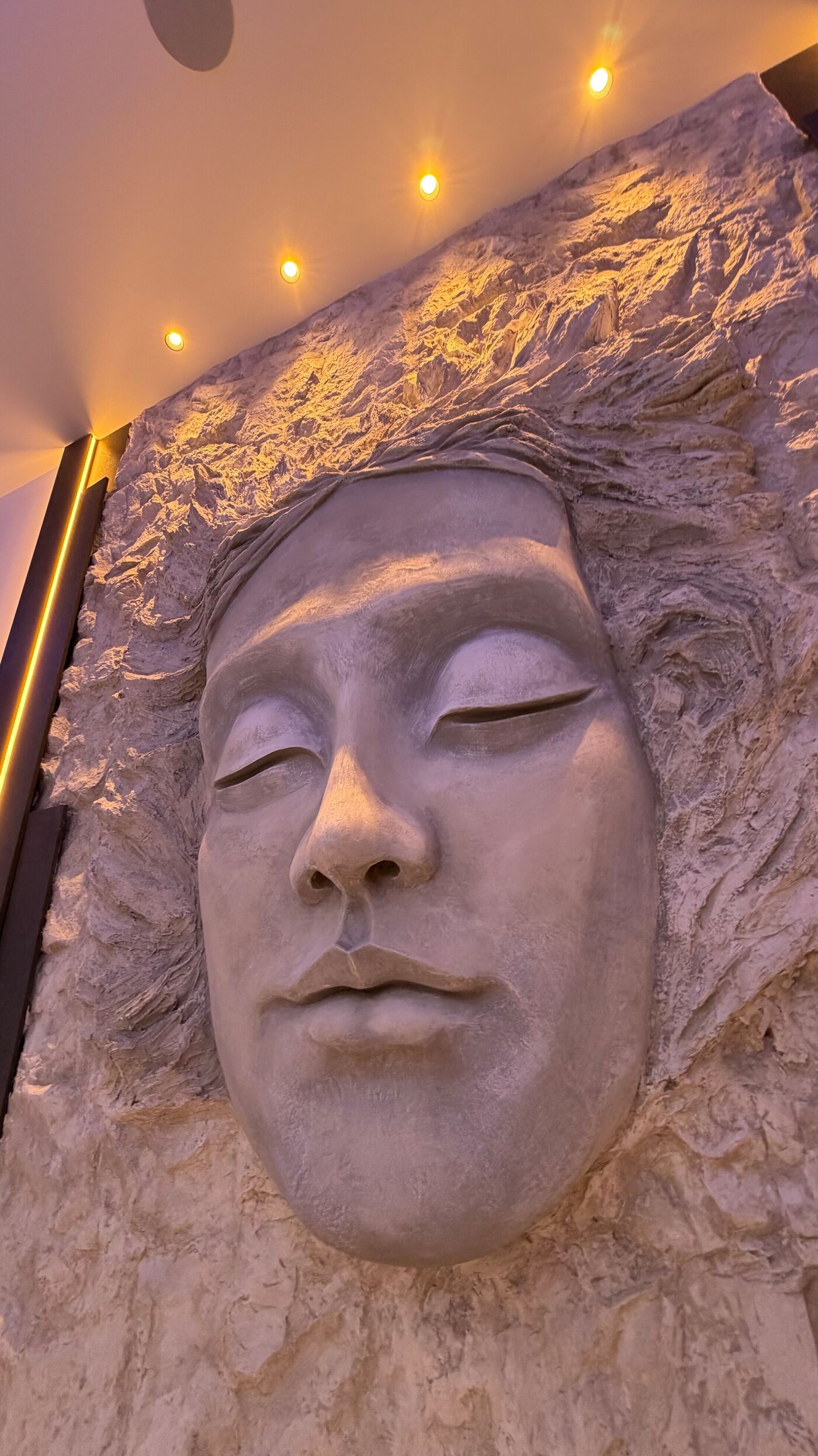 Face in relief with closed eyes emerging from a textured wall, lit by warm ceiling spots.