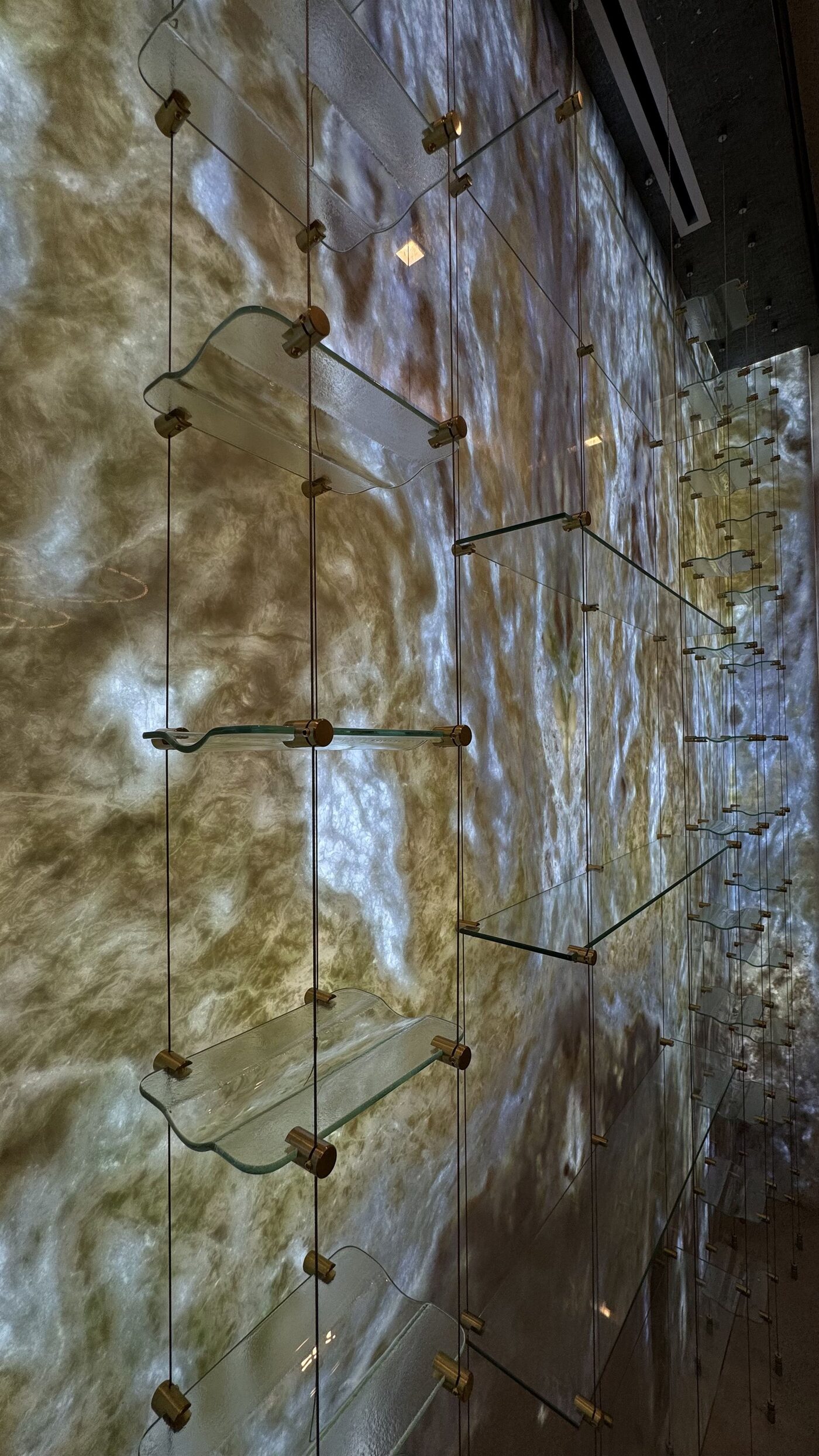Backlit onyx wall with suspended glass shelves held by bronze fittings.