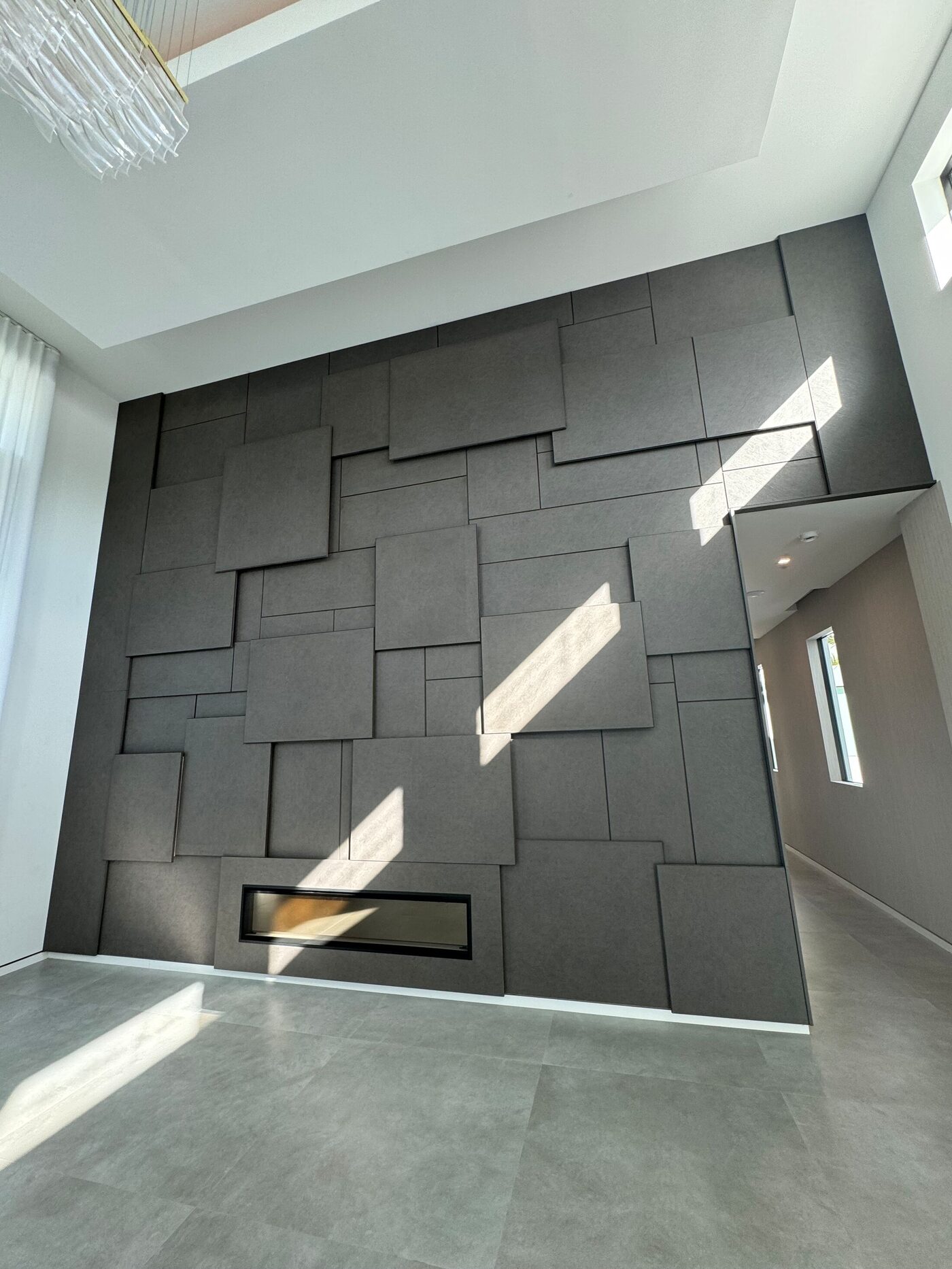 Full view of a double-height wall of dark panels with a linear fireplace and sunlight on the floor.