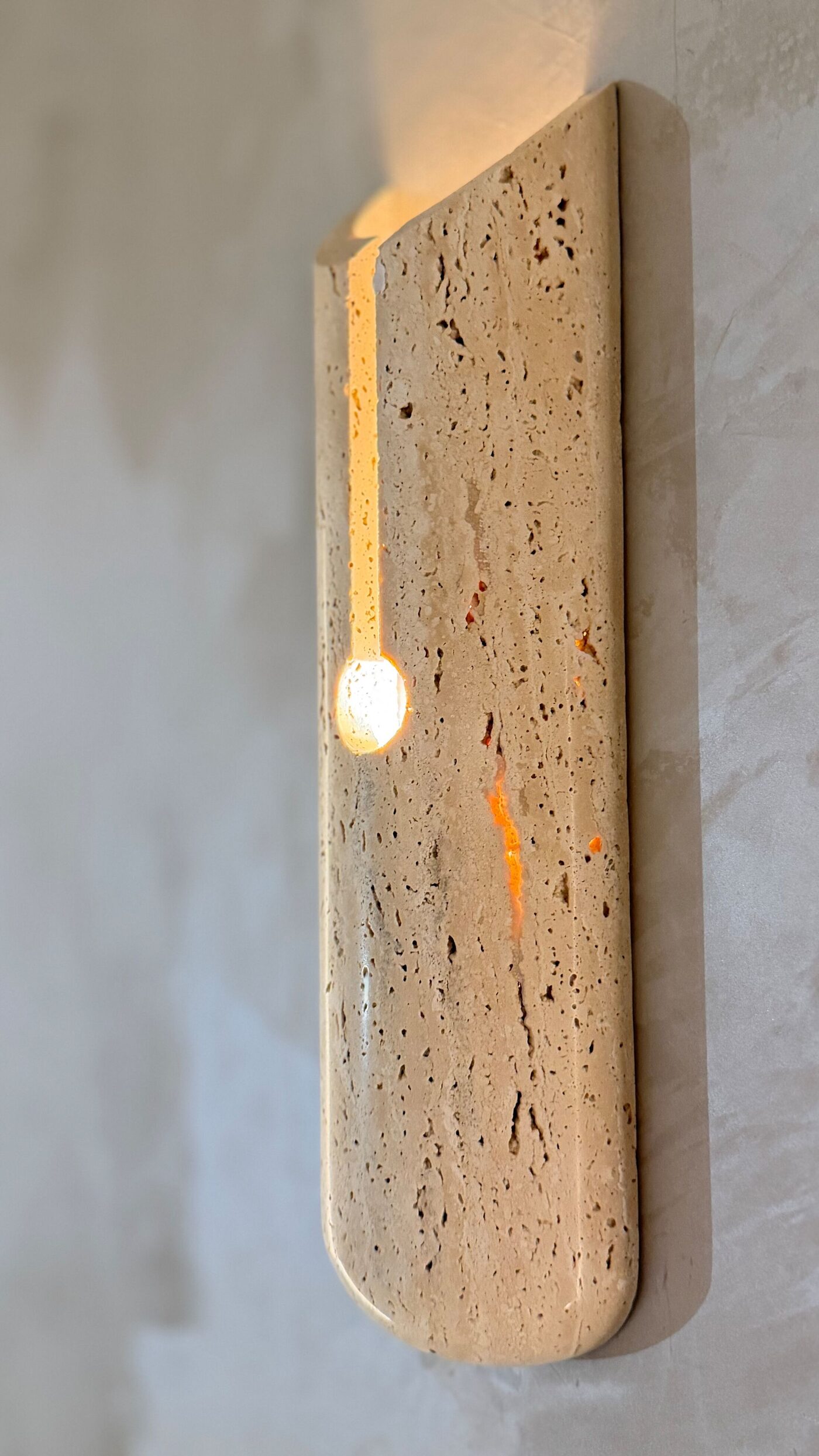 A luminaire carved in natural travertine on a textured wall, with warm LED light.