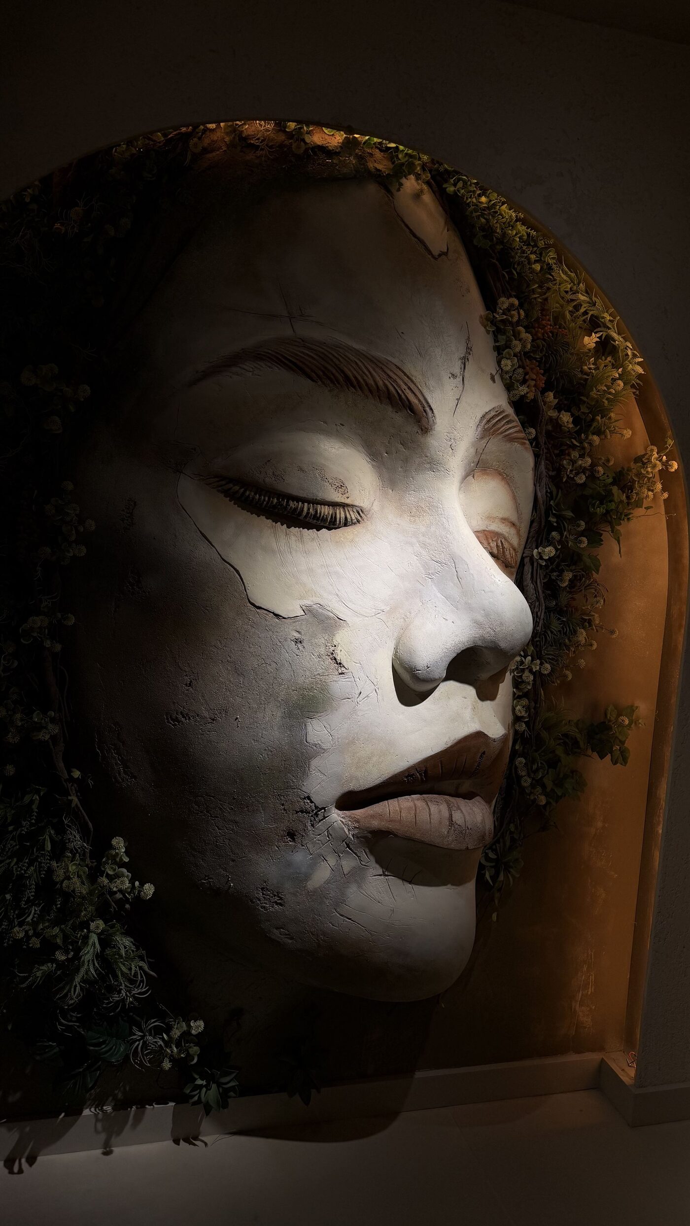Large sculpted face with closed eyes set inside an arched niche, framed by greenery and warm light.