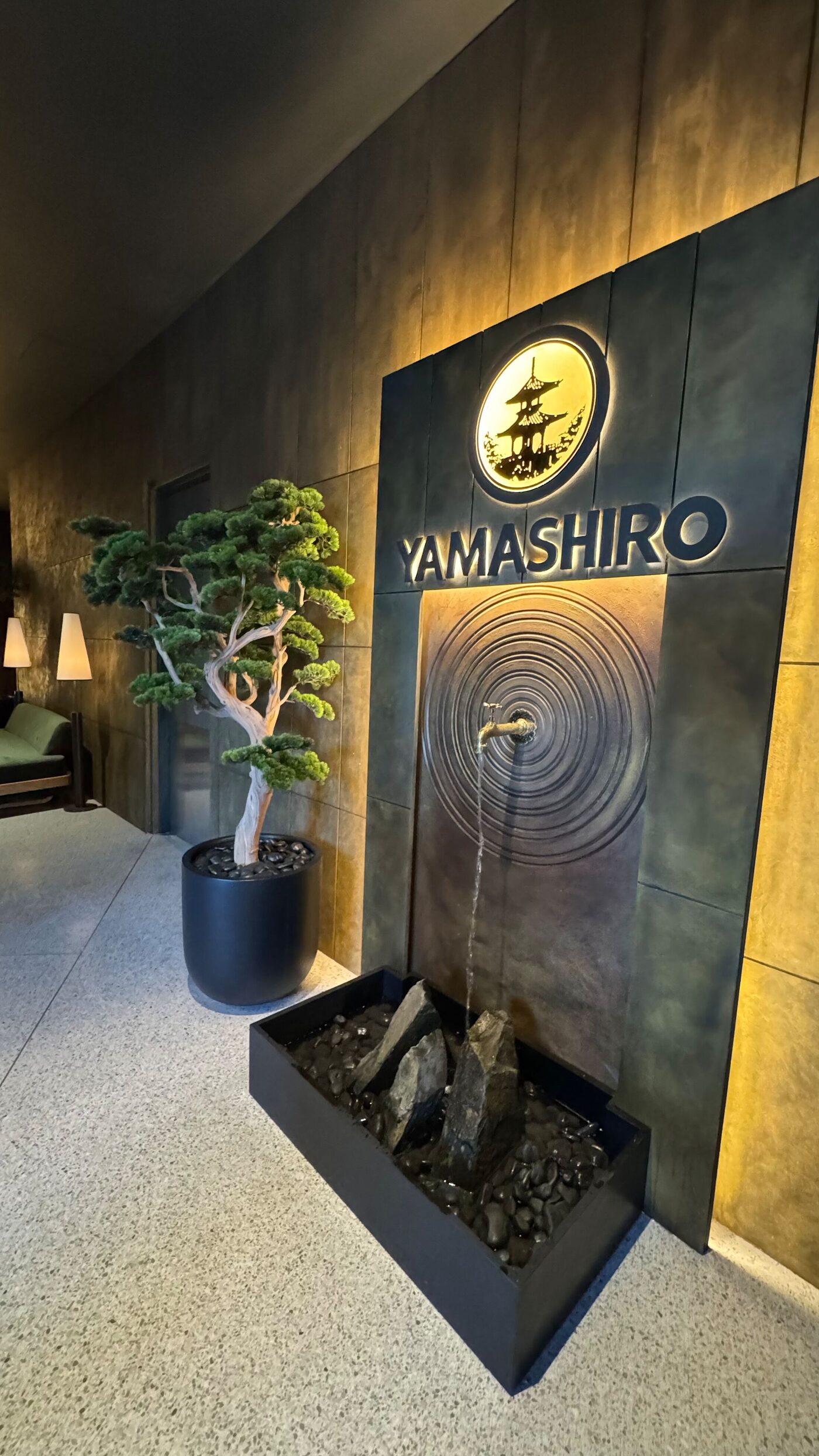Restaurant entrance with a dark backlit wall, the Yamashiro sign, a water fountain and a bonsai tree.