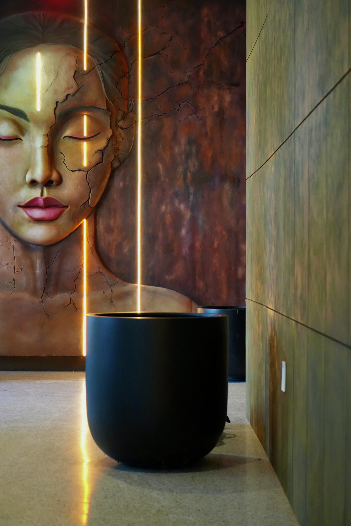 The painted face with closed eyes, crossed by vertical lines of warm LED light, with a black planter in front and patinated wall panels.